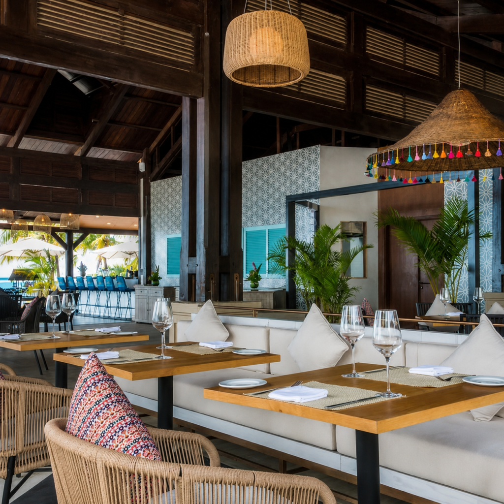 Restaurant tropical élégant avec mobilier en bois, tables dressées avec assiettes, serviettes blanches et verres à vin, banquettes crème, fauteuils en rotin, poutres en bois sombre, suspensions en fibres naturelles, murs décoratifs à motifs, volets turquoise, plantes tropicales et espace bar ouvert sur le lagon de l'ile aux Cerfs.
